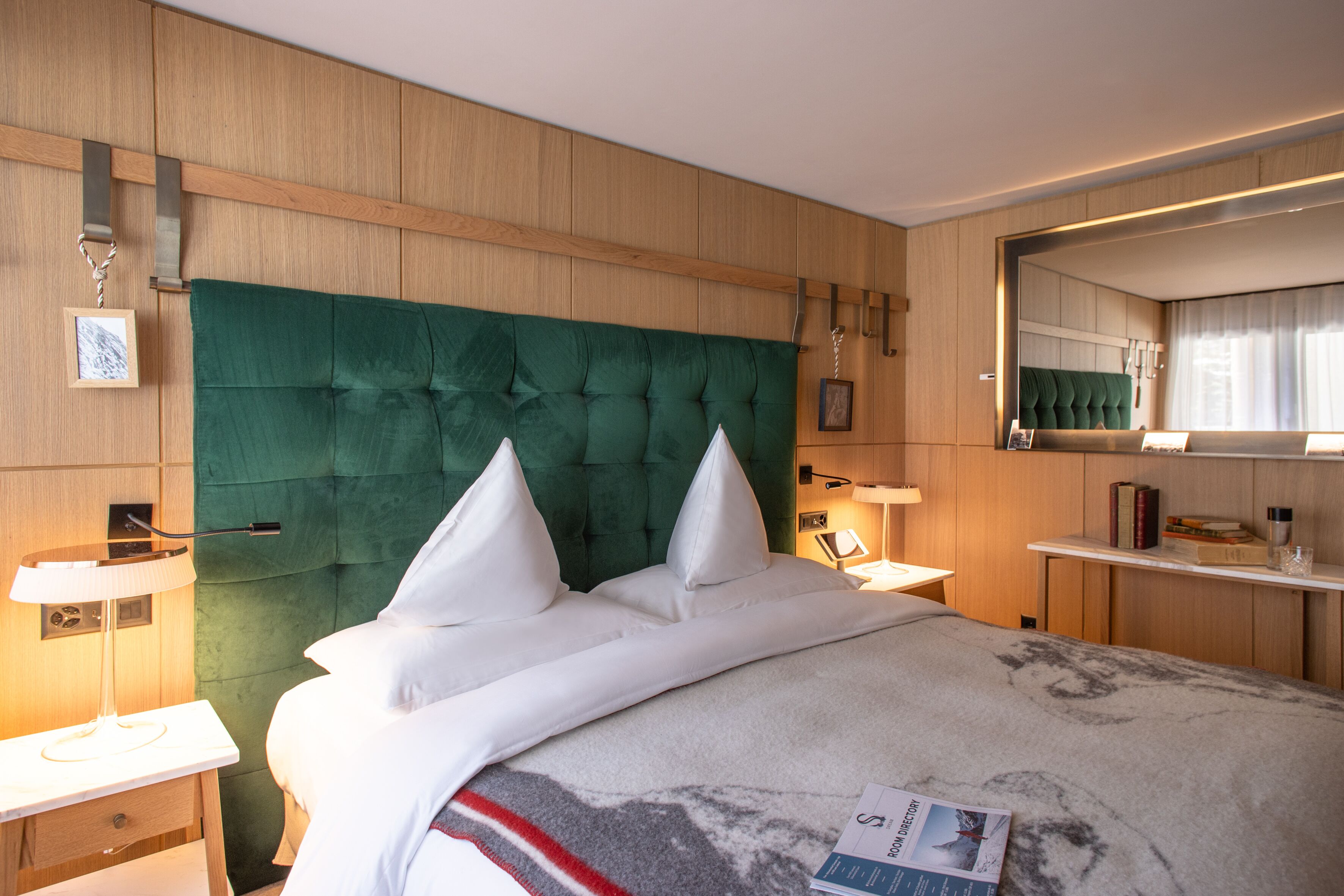 A hotel room with a large bed, a wooden headboard, bedside tables with lamps, a mirror, and a small shelf with books and a glass.