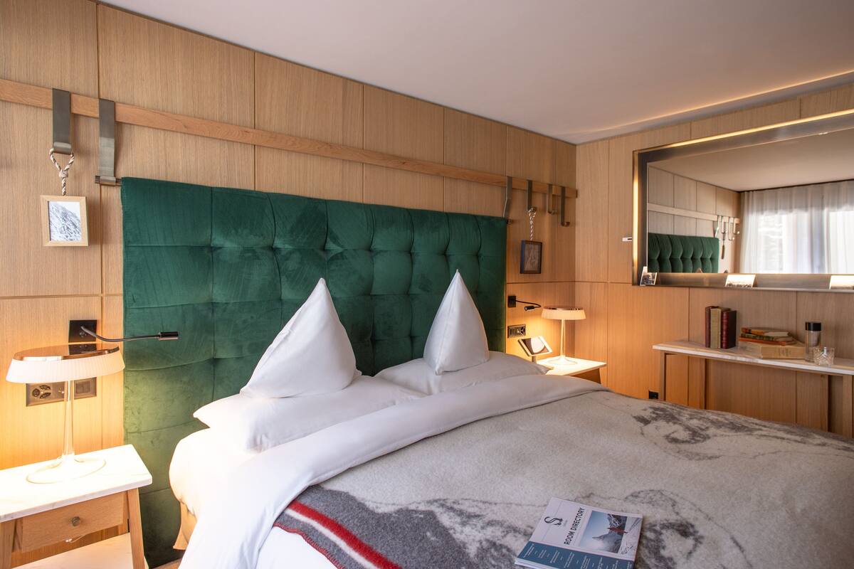 A hotel room with a large bed, a wooden headboard, bedside tables with lamps, a mirror, and a small shelf with books and a glass.