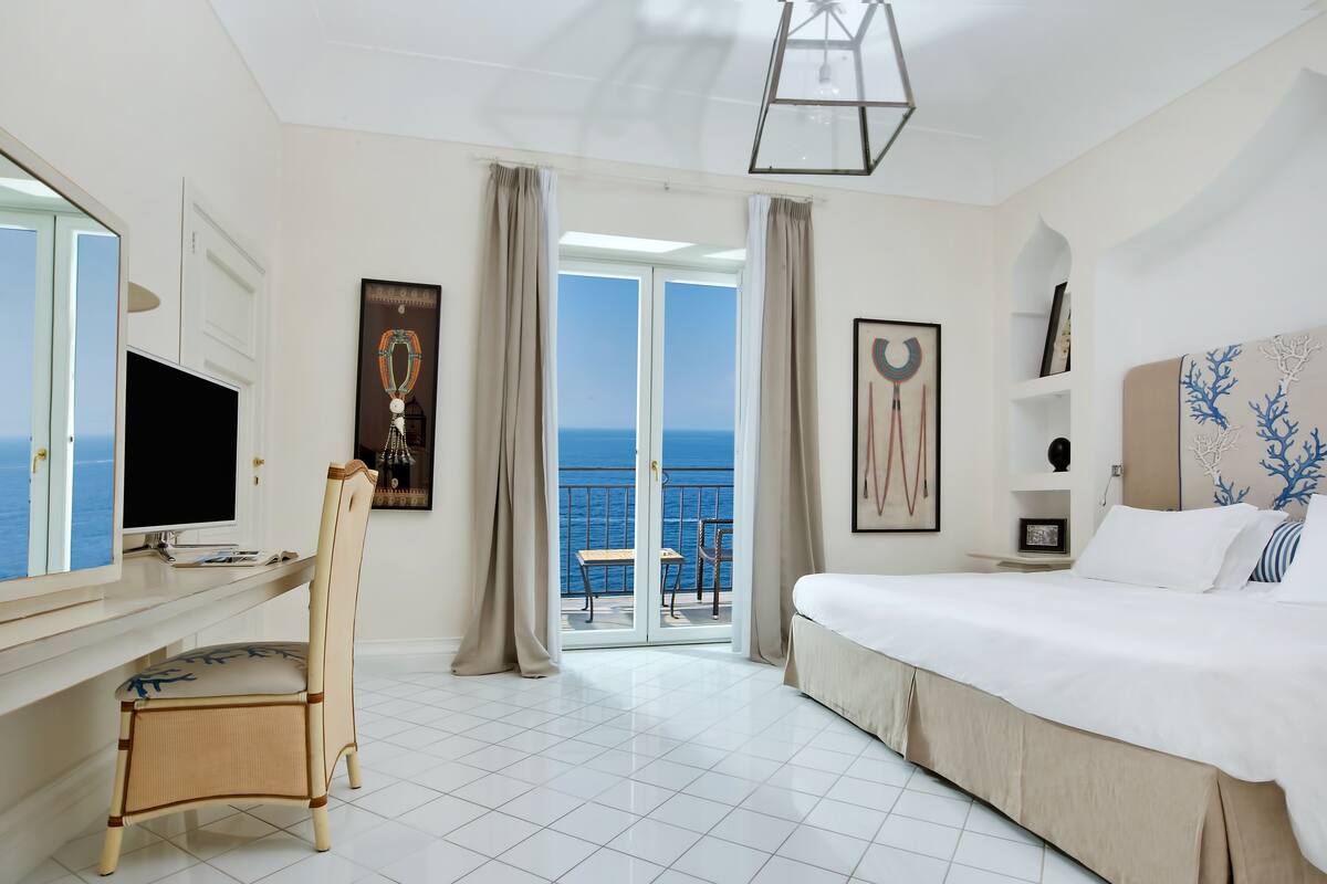 A hotel room with a bed, a desk with a computer, a chair, a TV, and a balcony with a view of the sea.