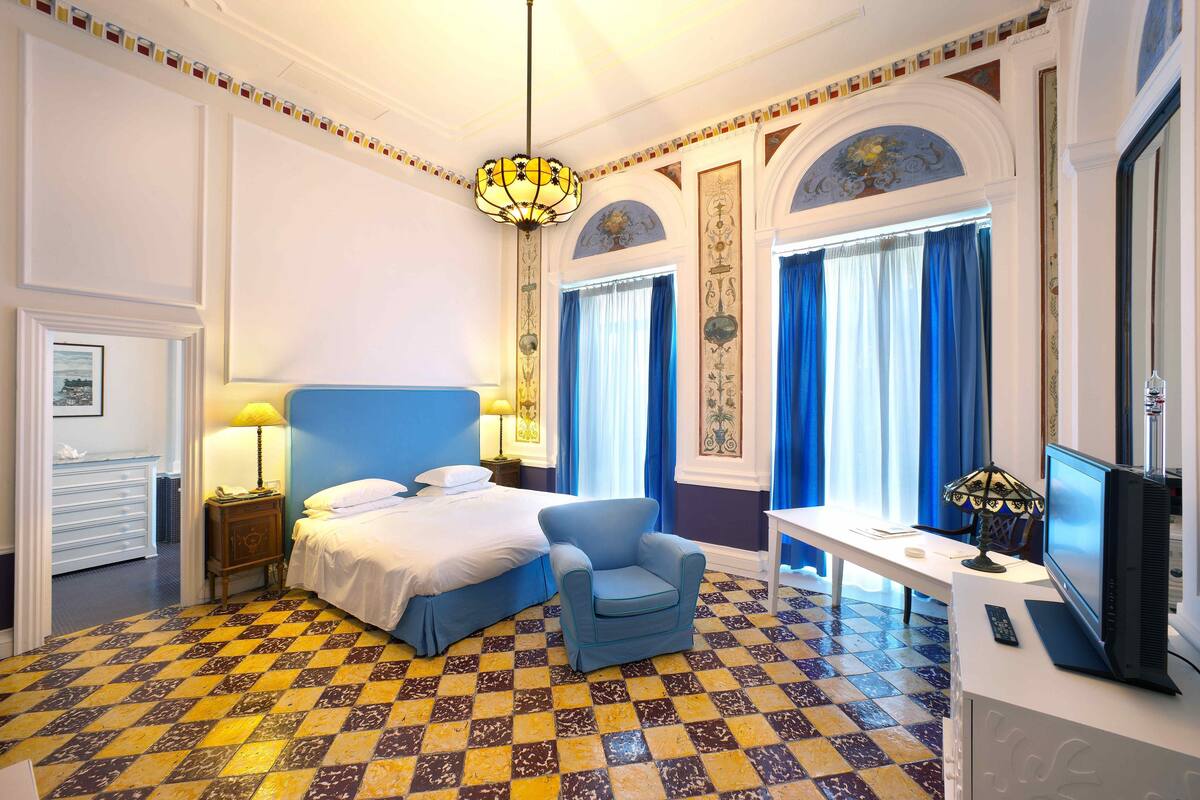 A hotel room with a checkered floor, a bed with blue bedding, a blue armchair, a desk with a computer, and a television.