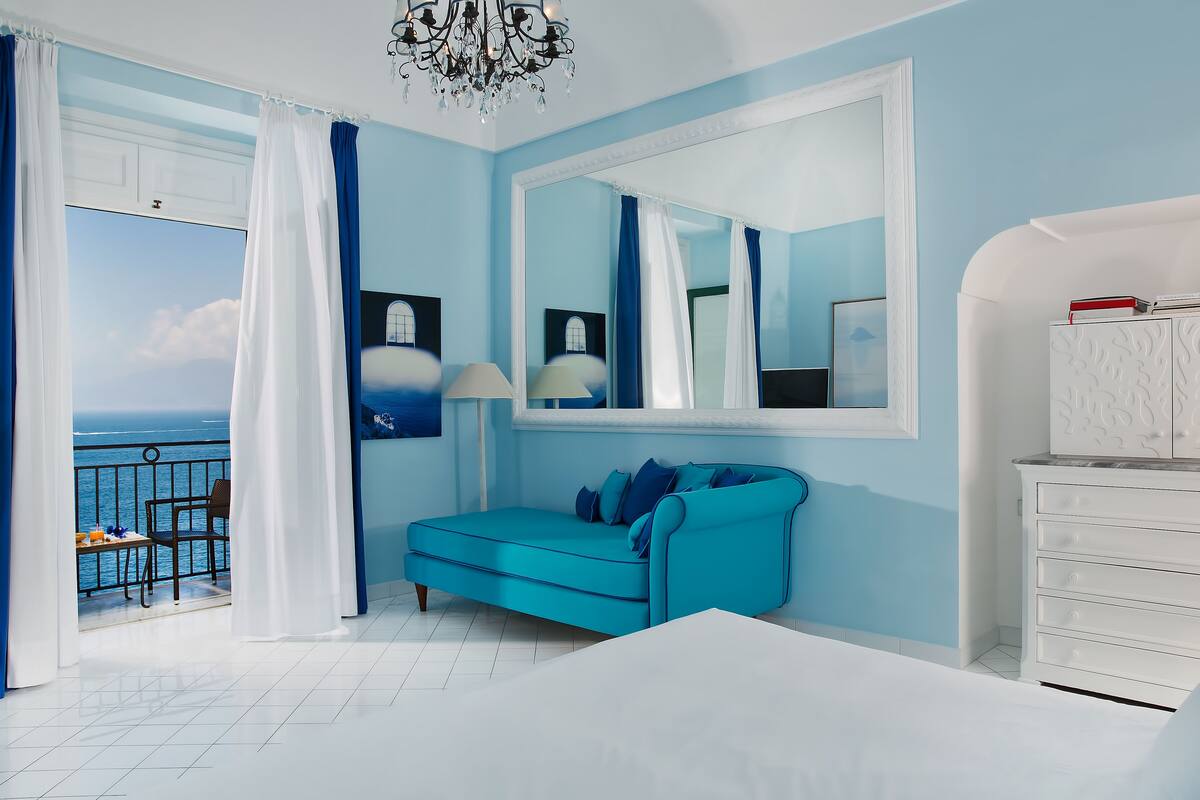 A bedroom with a large bed, a blue sofa, a white dresser, and a balcony with a view of the sea.