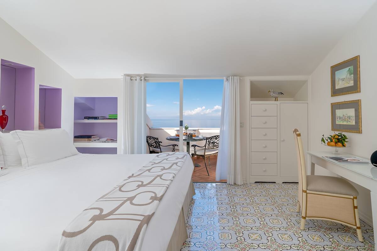 A hotel room with a bed, a desk, a chair, a bookshelf, and a balcony with a view of the sea.