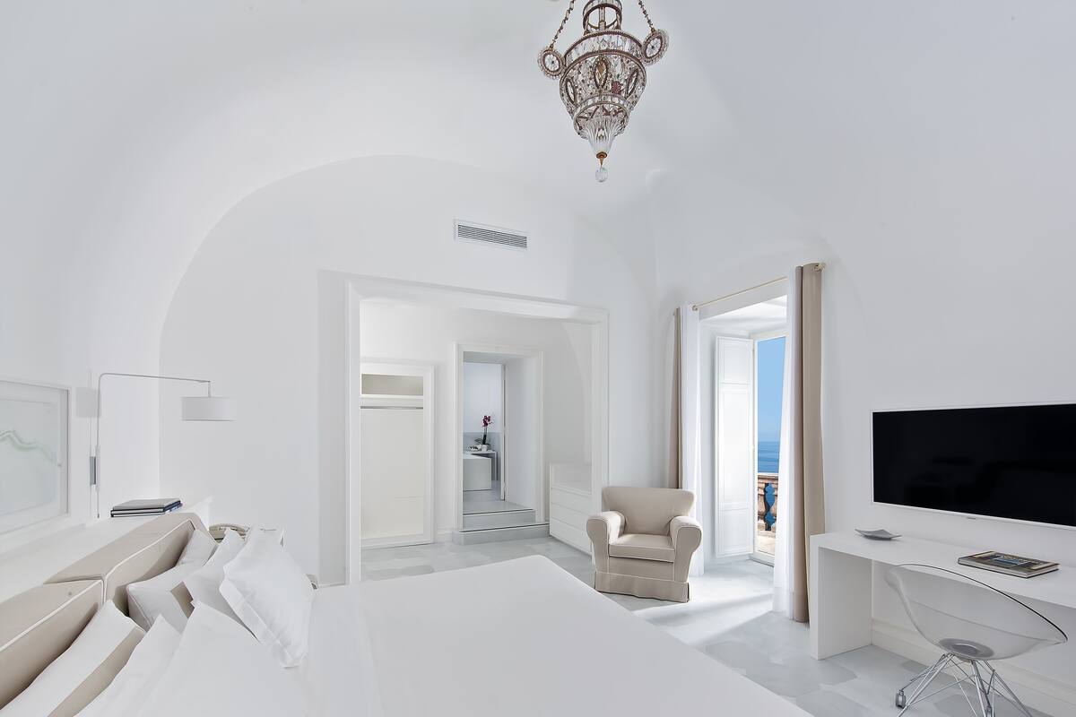 A modern hotel room with a large bed, a TV, a desk, and a chair. There is a chandelier and a view of the sea through a window.