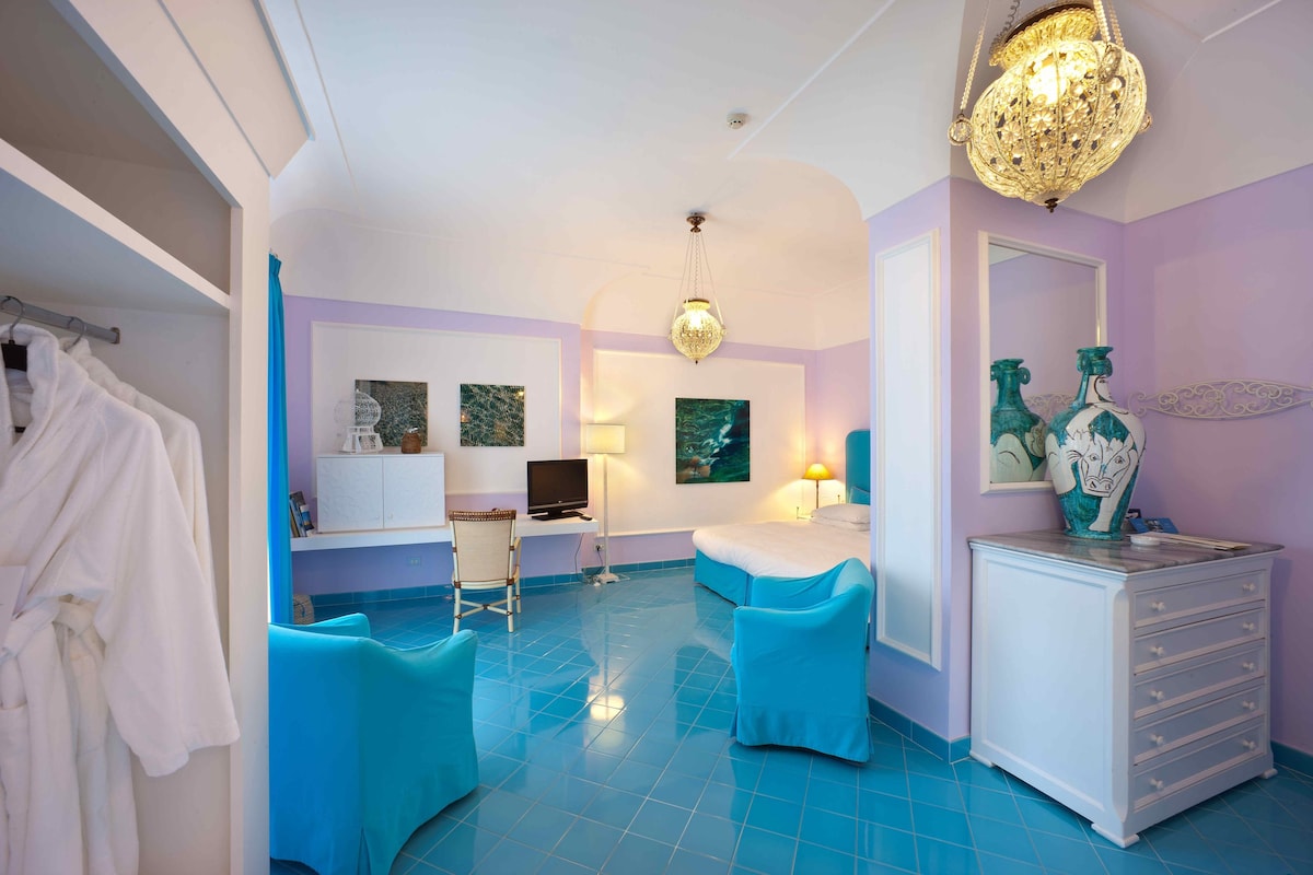 A hotel room with a blue tiled floor, a desk with a computer, a bed, two blue chairs, a white dresser, and a chandelier.