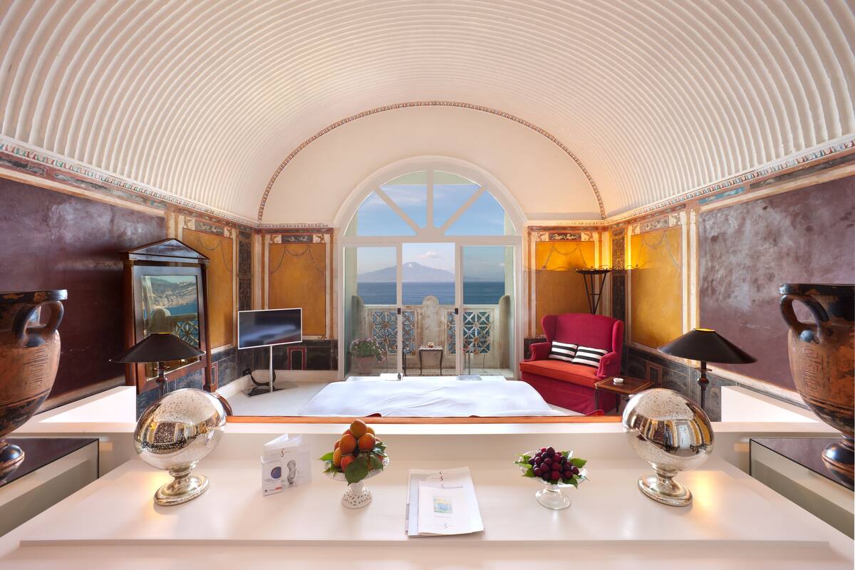 A luxurious room with a large arched ceiling, a bed, a desk with a lamp, and a view of the sea.
