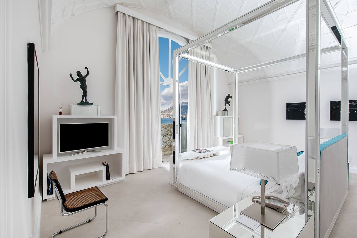 A modern hotel room with a large bed, a television, a desk with a lamp, and a view of the outdoors.