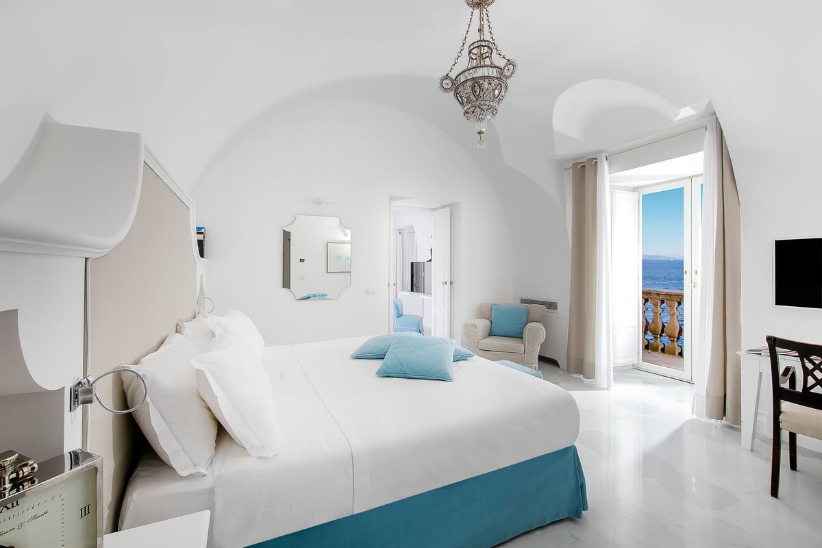 A modern hotel room with a large bed, a chandelier, a balcony with a view, and a TV.