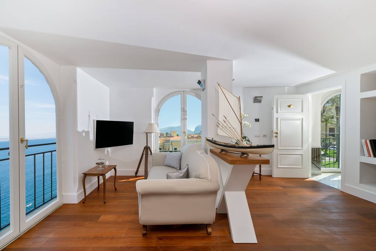 A spacious living area with a large window overlooking the sea, a comfortable sofa, a flat-screen TV, and a small table with a lamp.