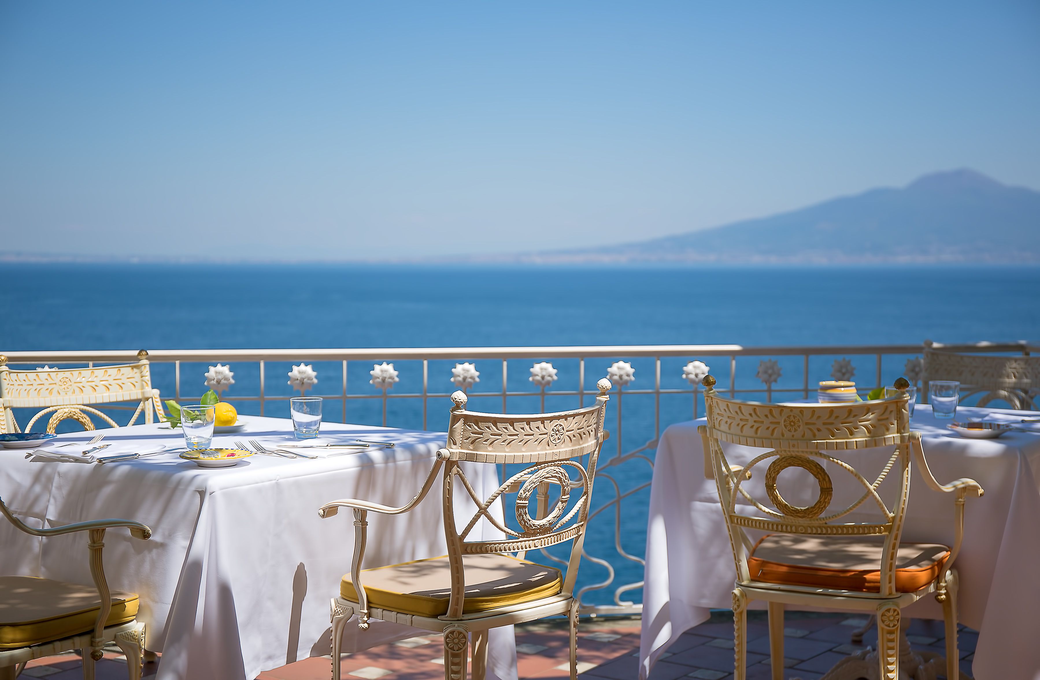 A terrace with white tables and chairs, a view of the sea, and a mountain in the background.