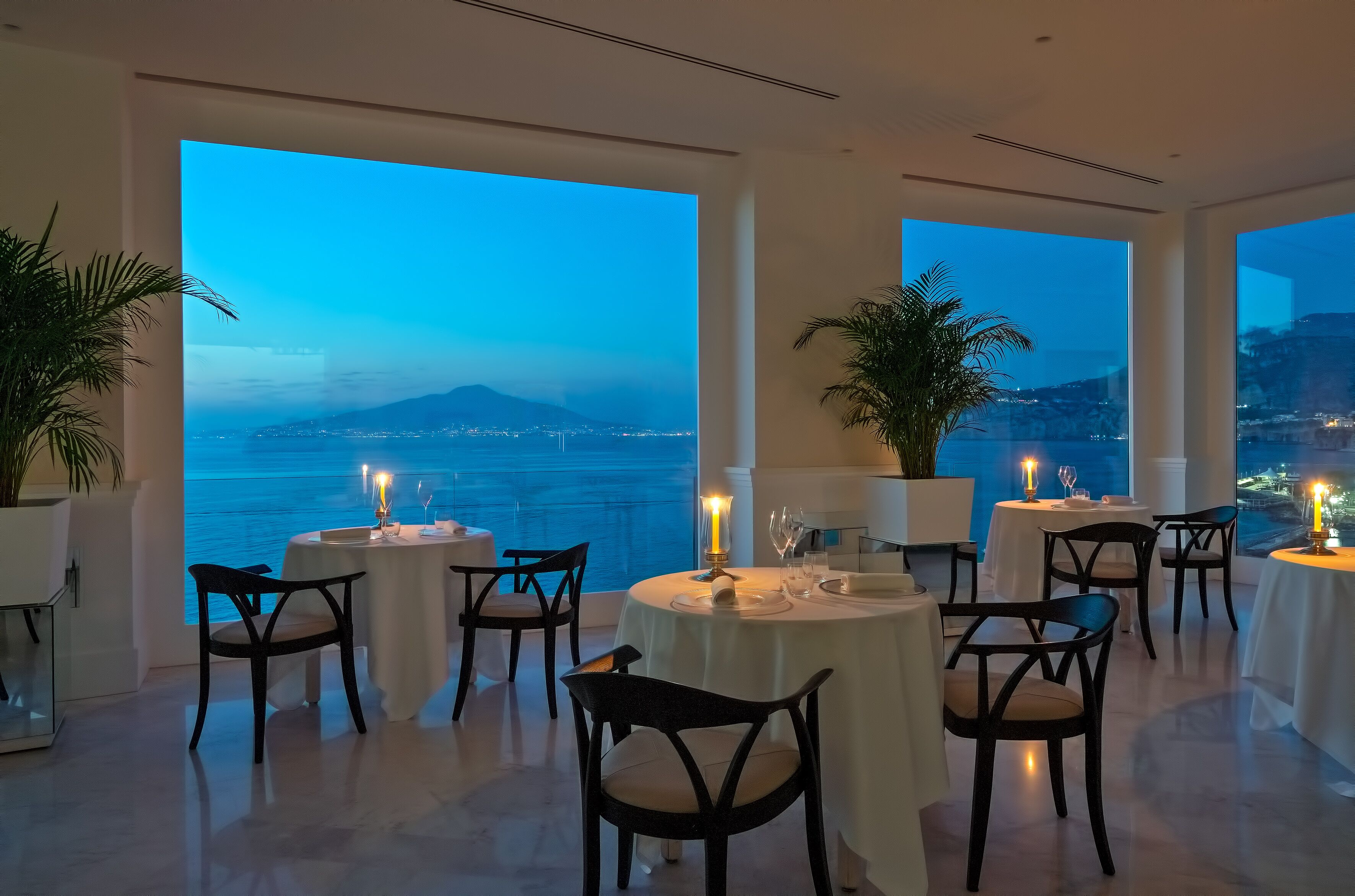 Un restaurante con mesas dispuestas para comer, amplias ventanas que ofrecen vista al mar y una montaña distante, y plantas en macetas.