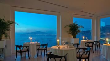 Un restaurante con mesas dispuestas para comer, amplias ventanas que ofrecen vista al mar y una montaña distante, y plantas en macetas.