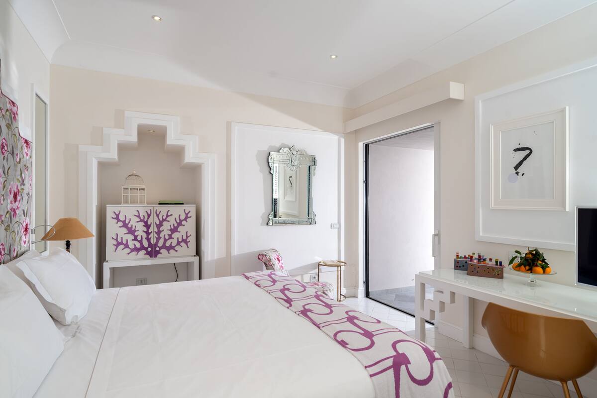 A modern hotel room with a bed, a desk, a chair, a mirror, and a door leading to another room.