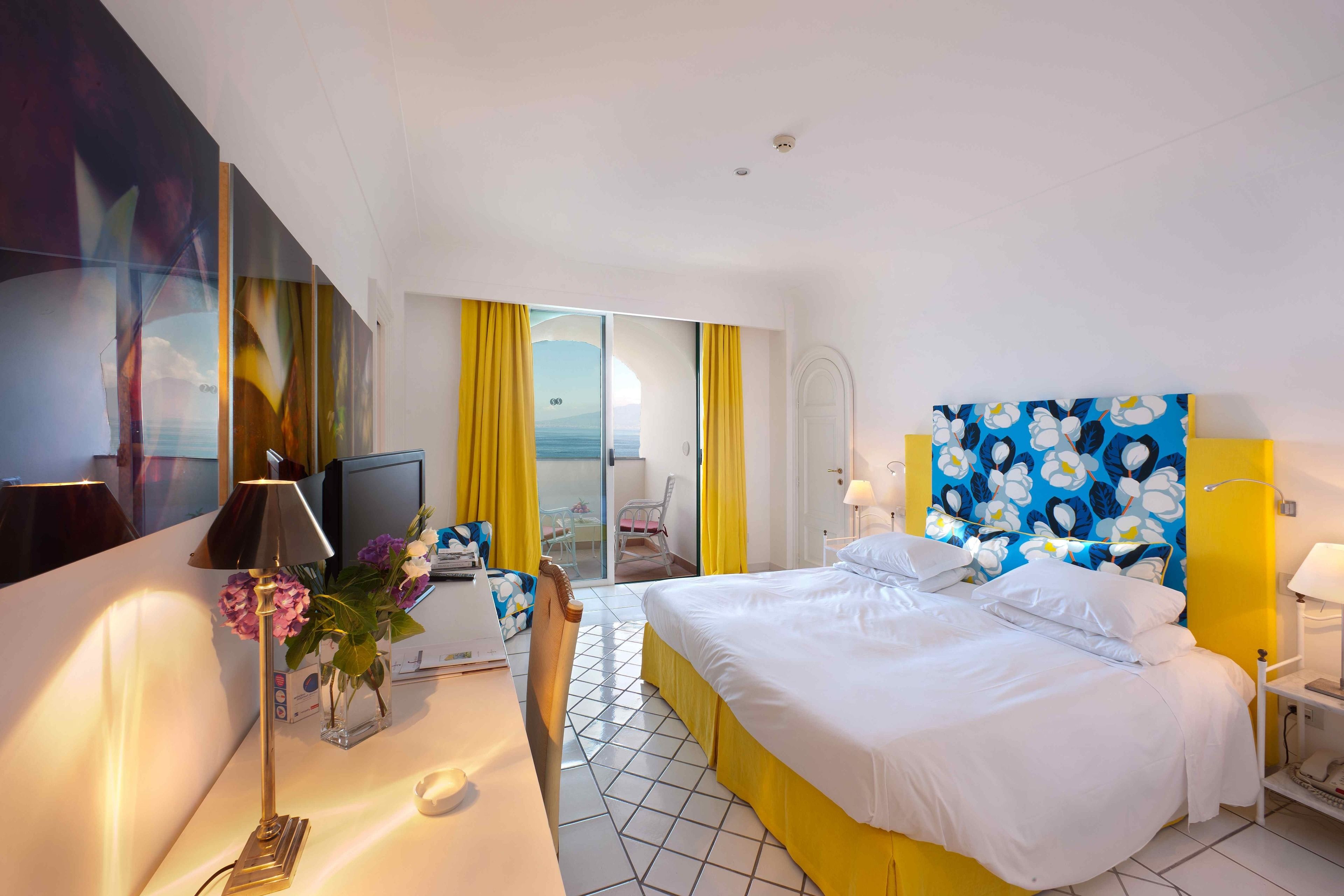 A hotel room with a bed, a desk, a chair, a TV, and a view of the sea through a sliding door.