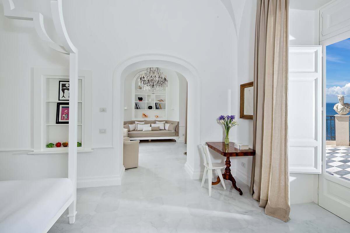 A spacious living area with a sofa, a dining table with chairs, a chandelier, and a view of the sea through an open door.
