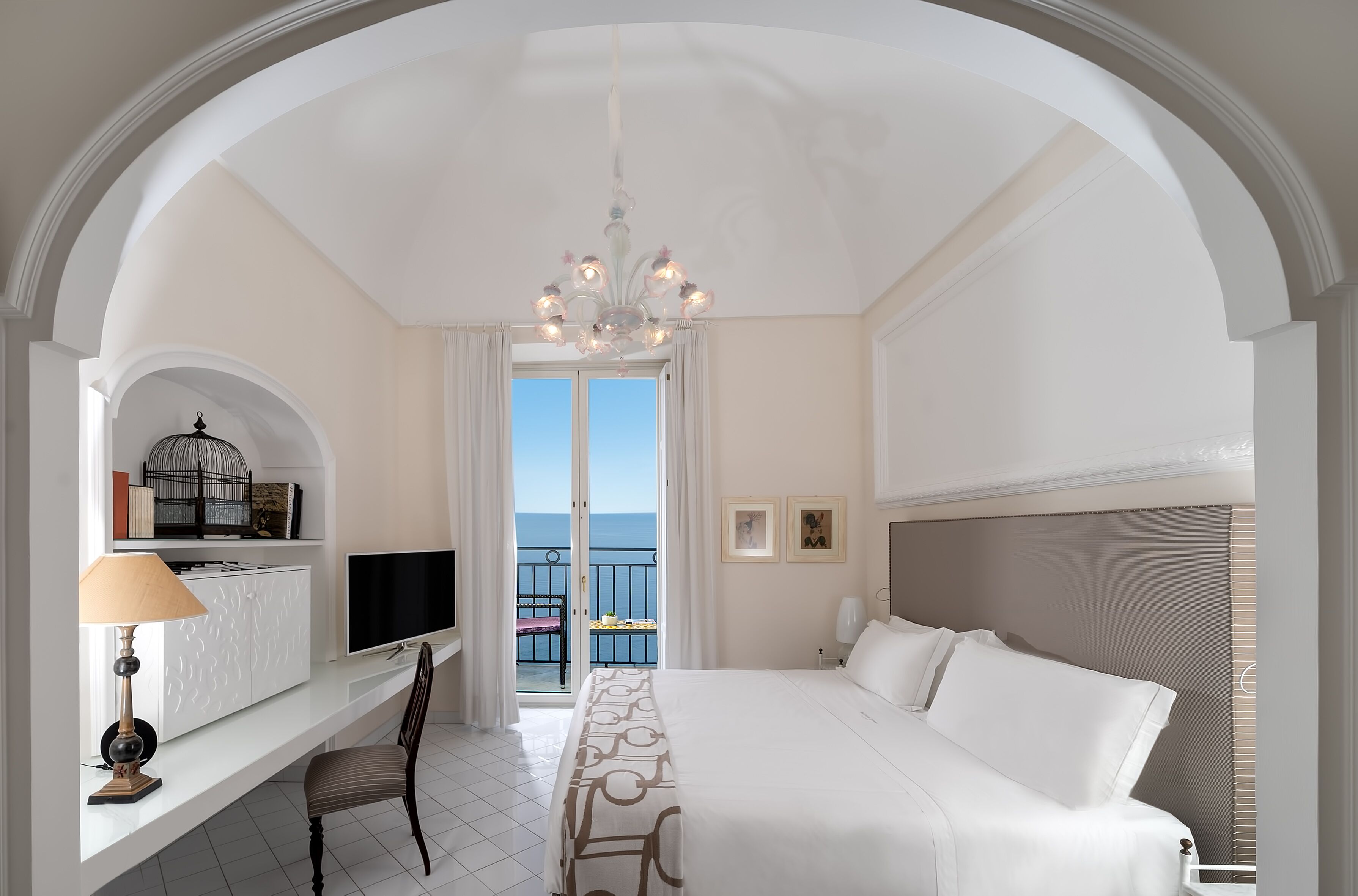 Habitación de hotel con una cama grande, un escritorio con televisor, una silla, balcón con vista al mar y un candelabro.
