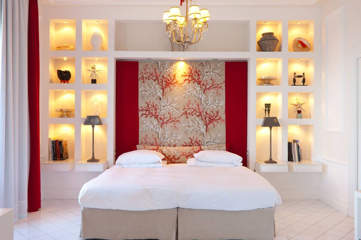 A modern bedroom with a large bed, built-in shelving with decorative items, a chandelier, and a unique coral-themed wall art.