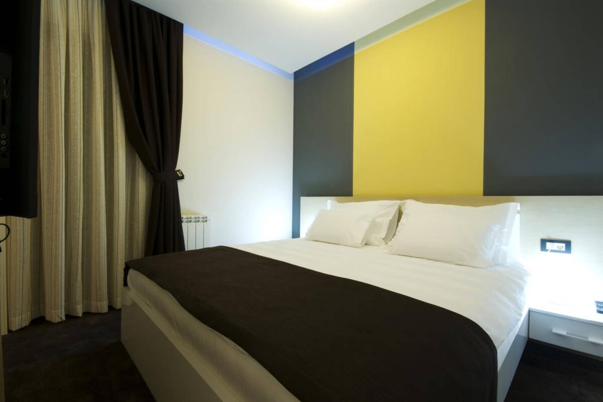 Een hotelkamer met een bed, gordijnen, een nachtkastje en een muur met een geometrisch ontwerp.