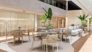 Un espace intérieur vaste et bien aménagé, avec différents types de sièges, notamment des tables rondes et des chaises, ainsi qu’un canapé. On y trouve également plusieurs plantes en pot et une grande fenêtre qui laisse entrer la lumière naturelle.