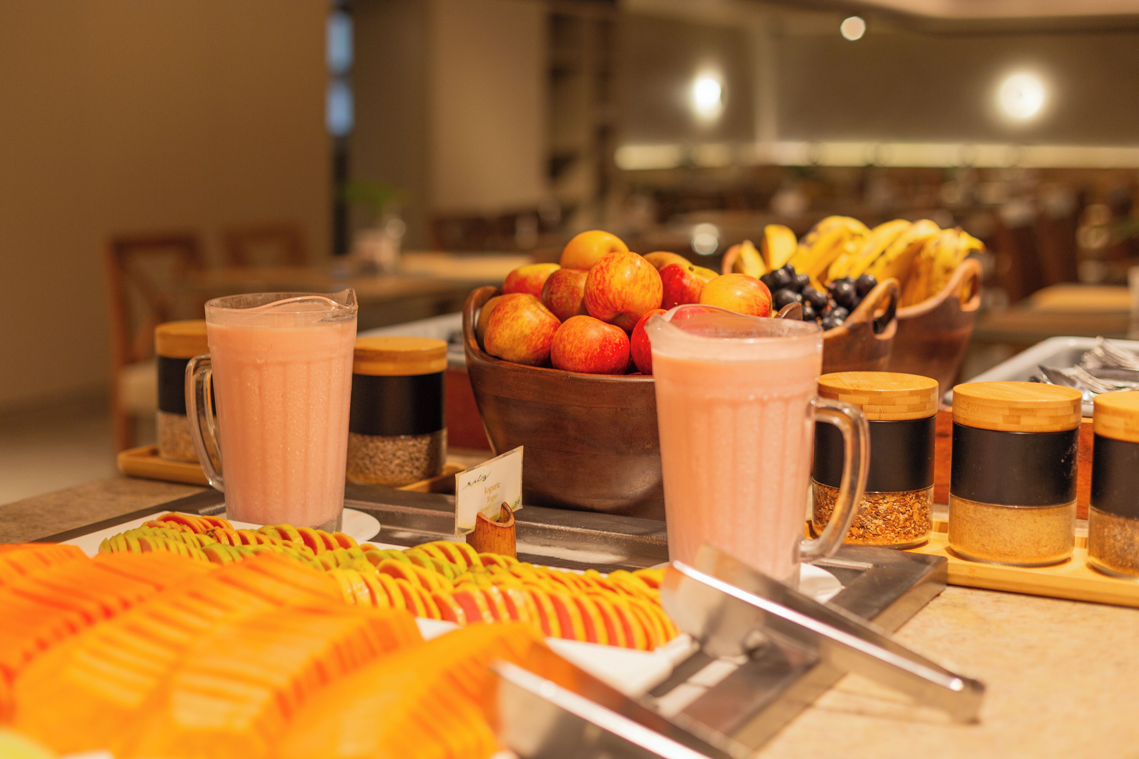 A buffet with various fruits, a bowl of oranges, and several mugs of a pink beverage.