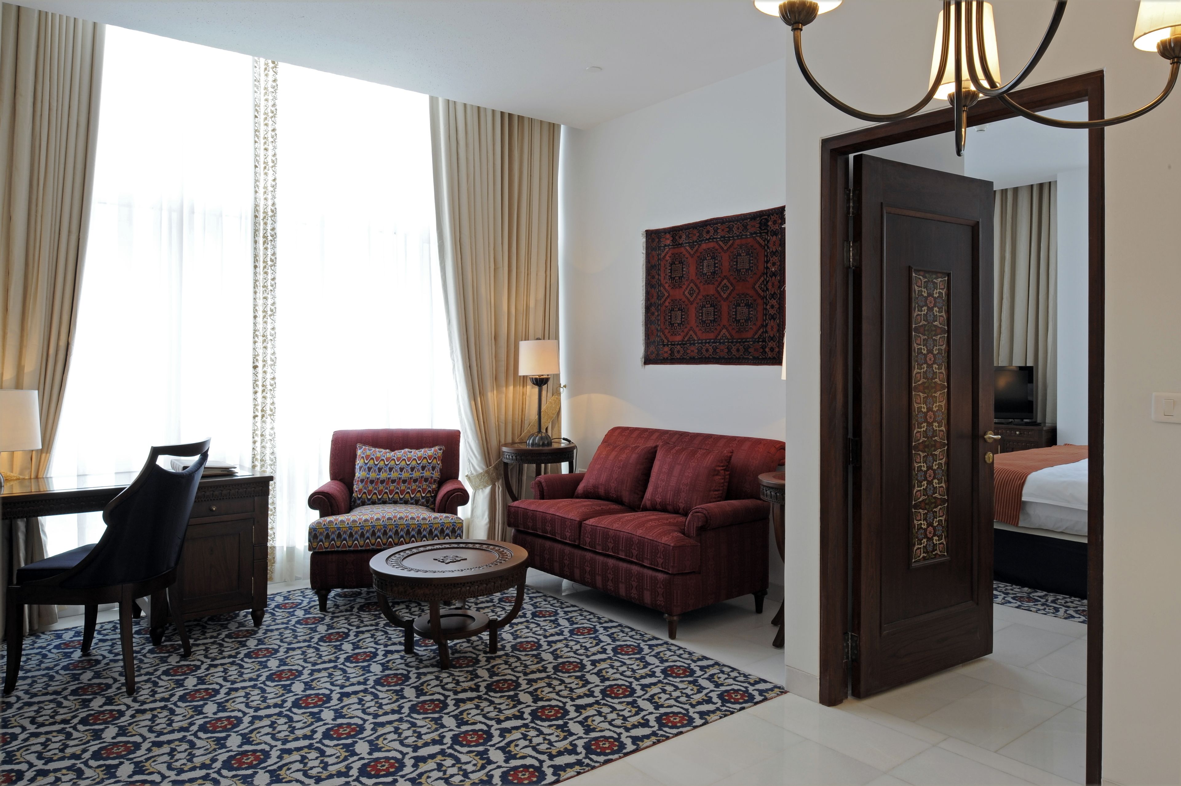 Habitación de hotel con sofá rojo, alfombra estampada, escritorio con silla y cama con cabecero decorado.