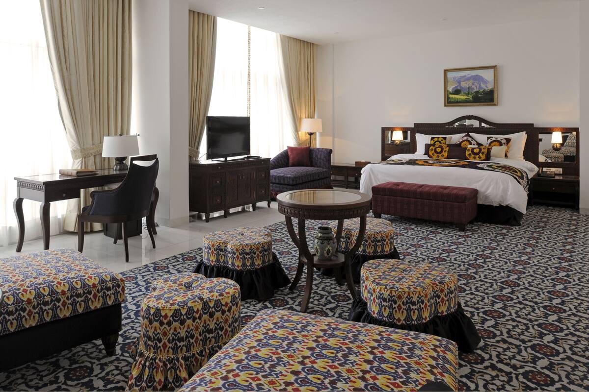 A hotel room with a bed, a desk, a chair, a TV, and a table with ottomans.