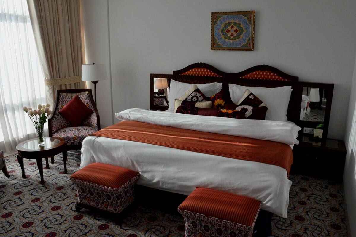 A hotel room with a large bed, two matching ottomans, a nightstand with a lamp, a chair, and a decorative mirror.