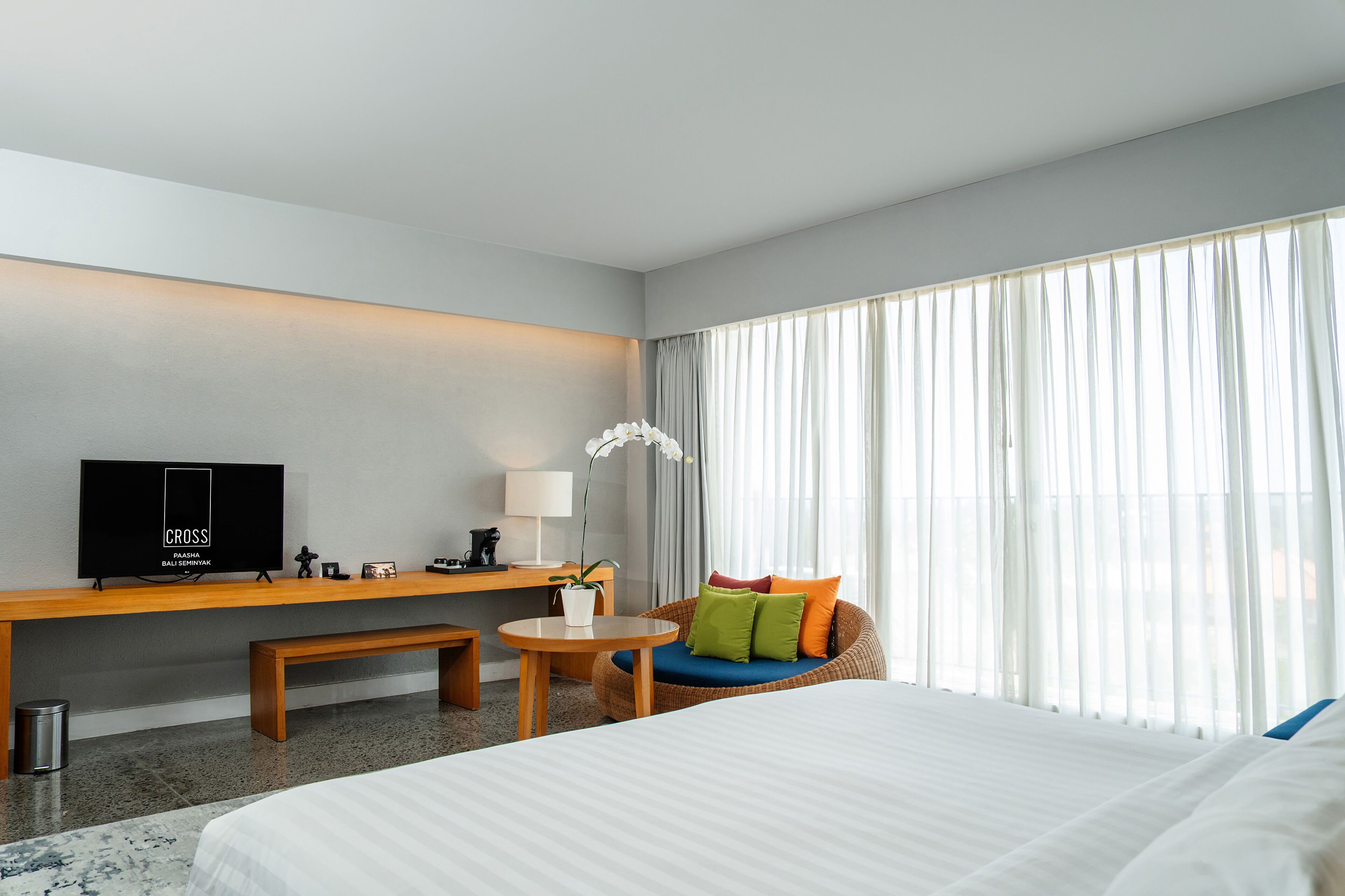 Habitación de hotel con cama, escritorio con televisor, mesita pequeña, banco y ventana con cortinas.