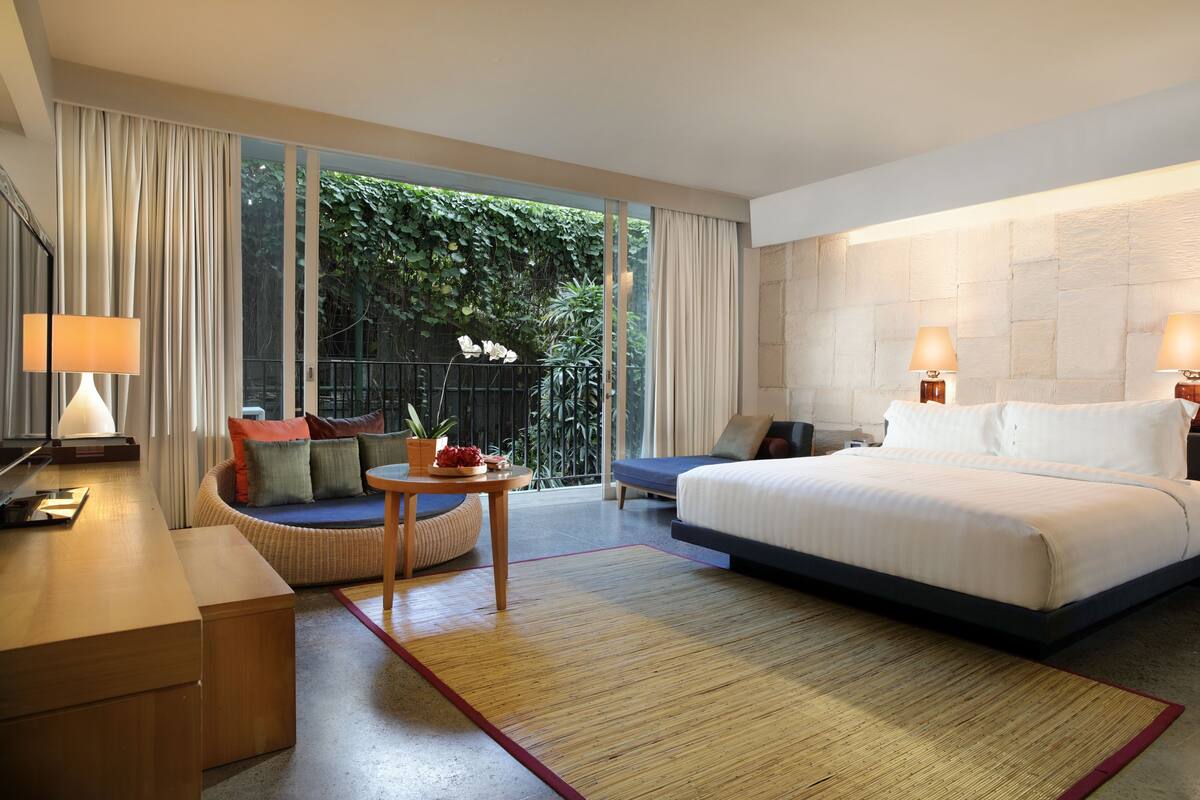 A modern hotel room with a large bed, a sofa, a small table with a plant, and a view of lush greenery through large windows.