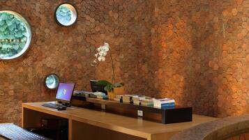 Mostrador de recepción con una computadora portátil, libros y una planta en maceta. Dos espejos redondos están sobre el mostrador y un banco a la izquierda. La pared de fondo está revestida con baldosas hexagonales.