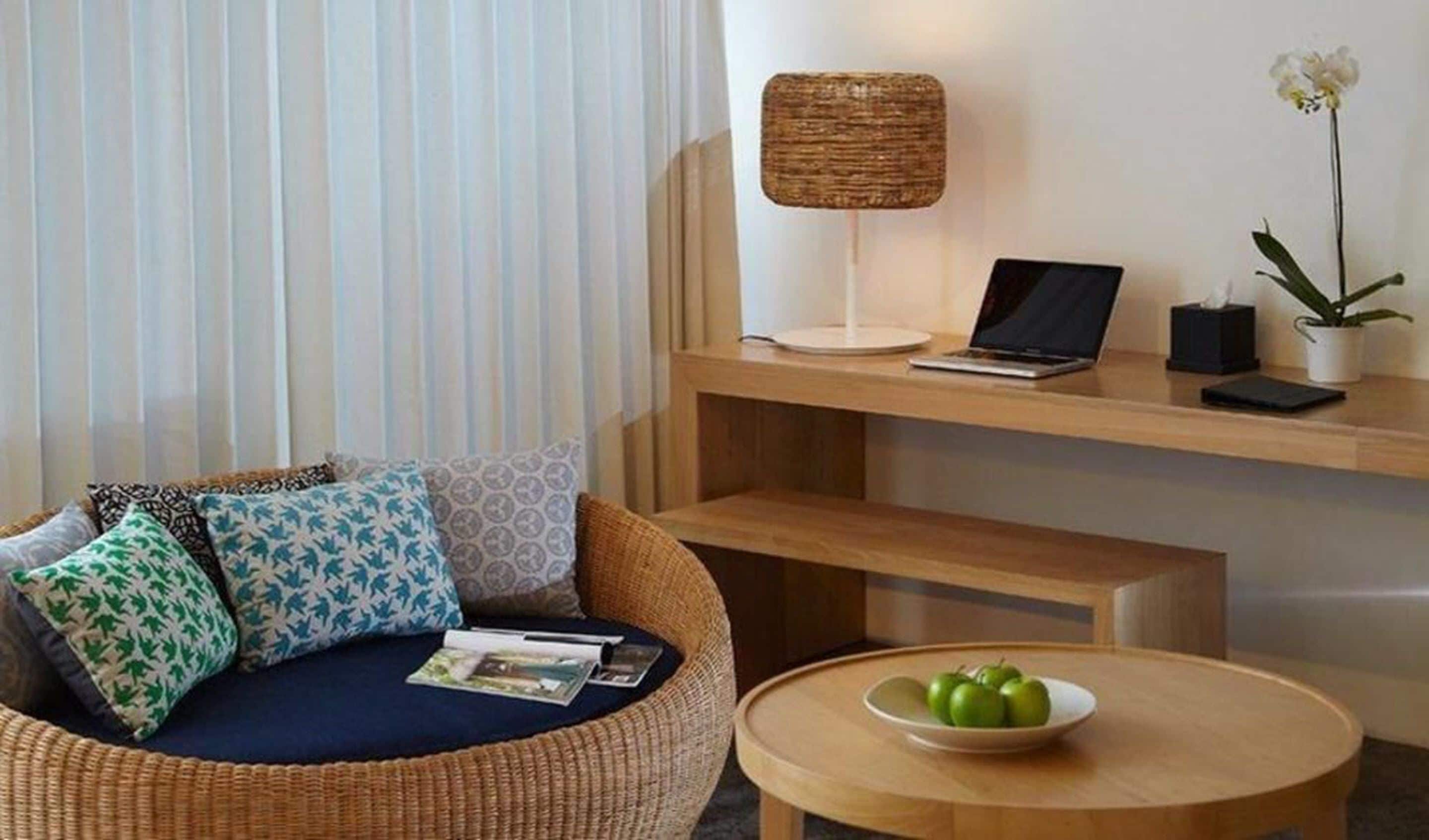 Ein modernes Wohnzimmer mit einem Rattan-Sofa, einem runden Holztisch, einem Laptop auf einem Holzregal und einer Topfpflanze.