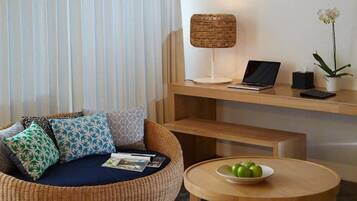 Ein modernes Wohnzimmer mit einem Rattan-Sofa, einem runden Holztisch, einem Laptop auf einem Holzregal und einer Topfpflanze.