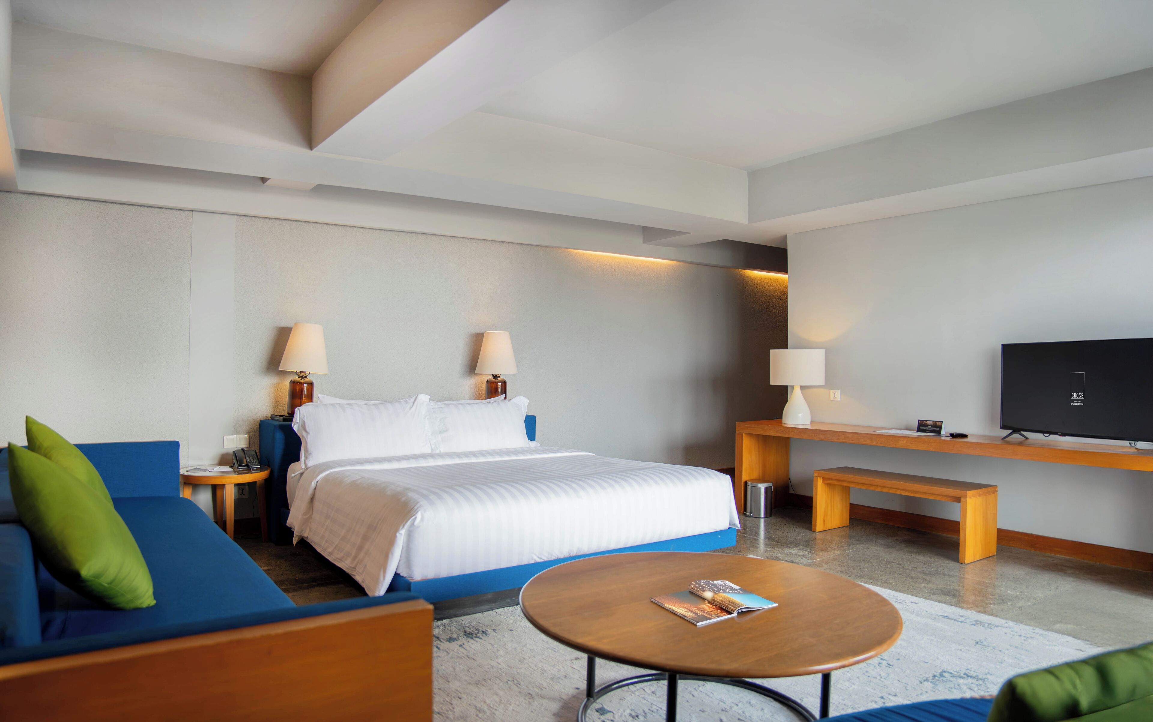 Una habitación de hotel moderna con cama, sofá azul, una pequeña mesa redonda, televisor y dos lámparas de noche.