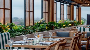 A restaurant with outdoor seating, tables set with glasses and cutlery, and large windows with vertical blinds. - Cross Paasha Bali Seminyak (Seminyak)