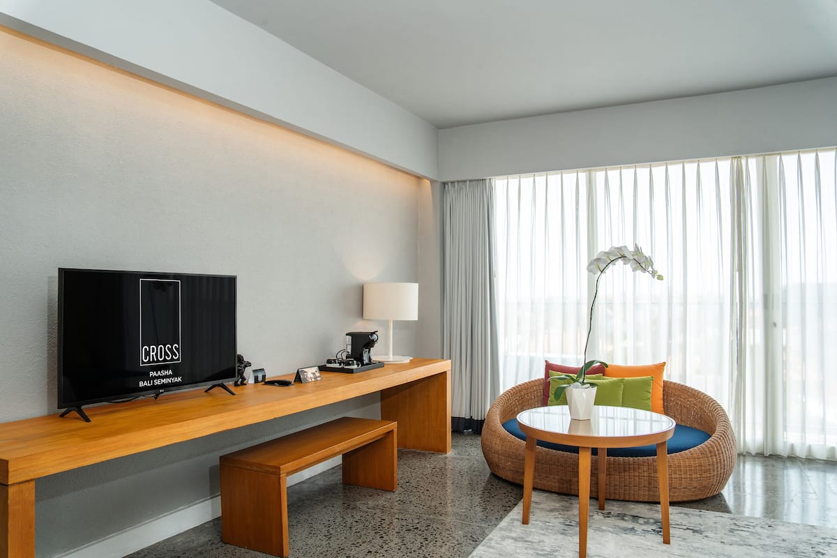 A modern hotel room with a wooden desk, a television, a lamp, a wicker sofa, and a small table with a plant.
