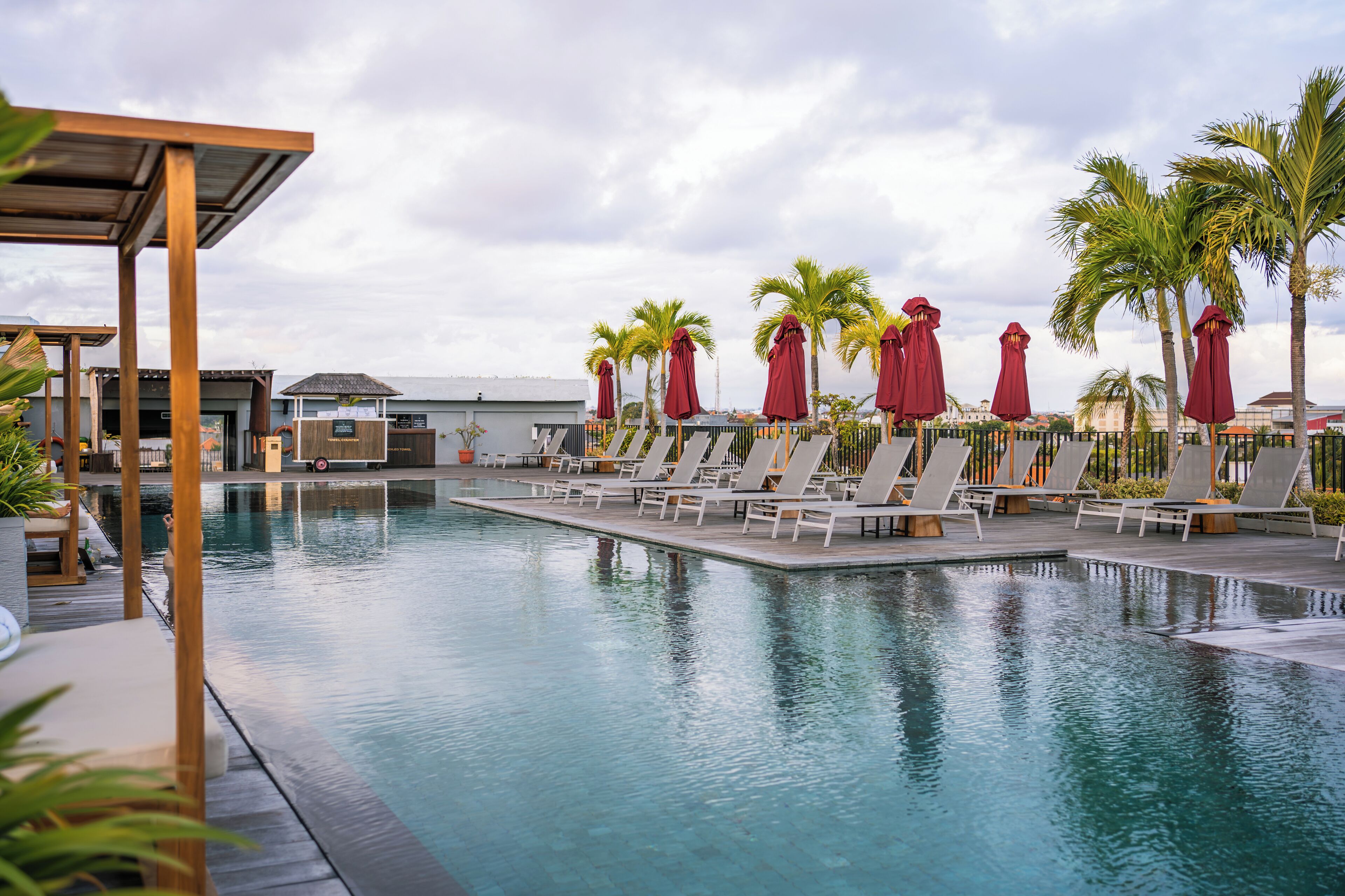 Piscina com cadeiras de descanso e guarda-sóis, cercada por palmeiras e um deck de madeira.