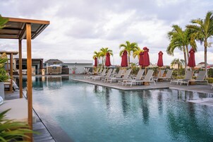 Piscina com cadeiras de descanso e guarda-sóis, cercada por palmeiras e um deck de madeira.
