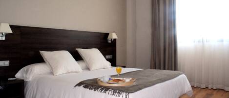 A neatly made bed with a tray of breakfast items, a bedside lamp, and a view of the outdoors through a window with curtains.
