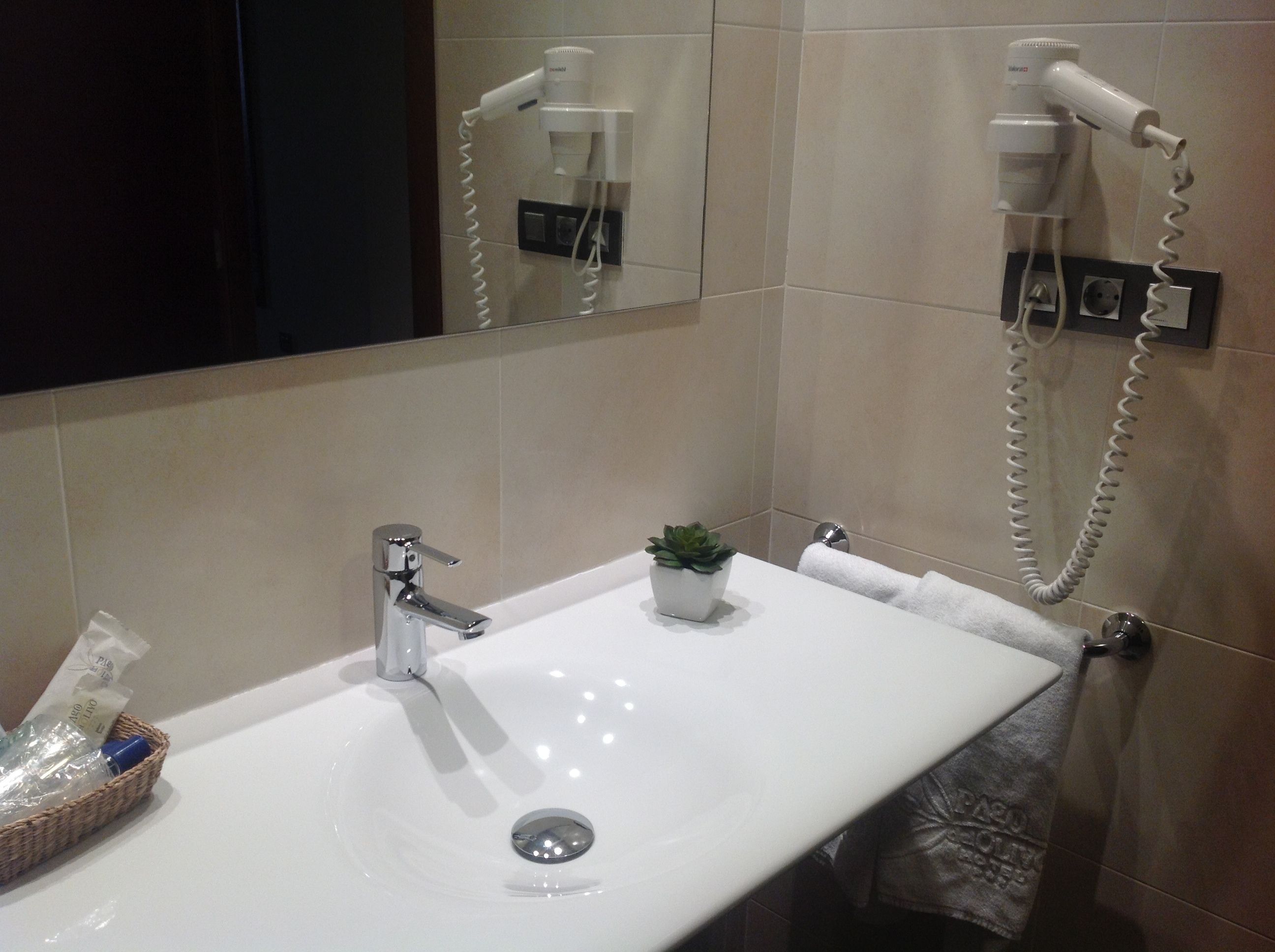 Un baño con un lavamanos blanco, una pequeña planta en maceta, un perchero y un secador de pelo sujeto a la pared.