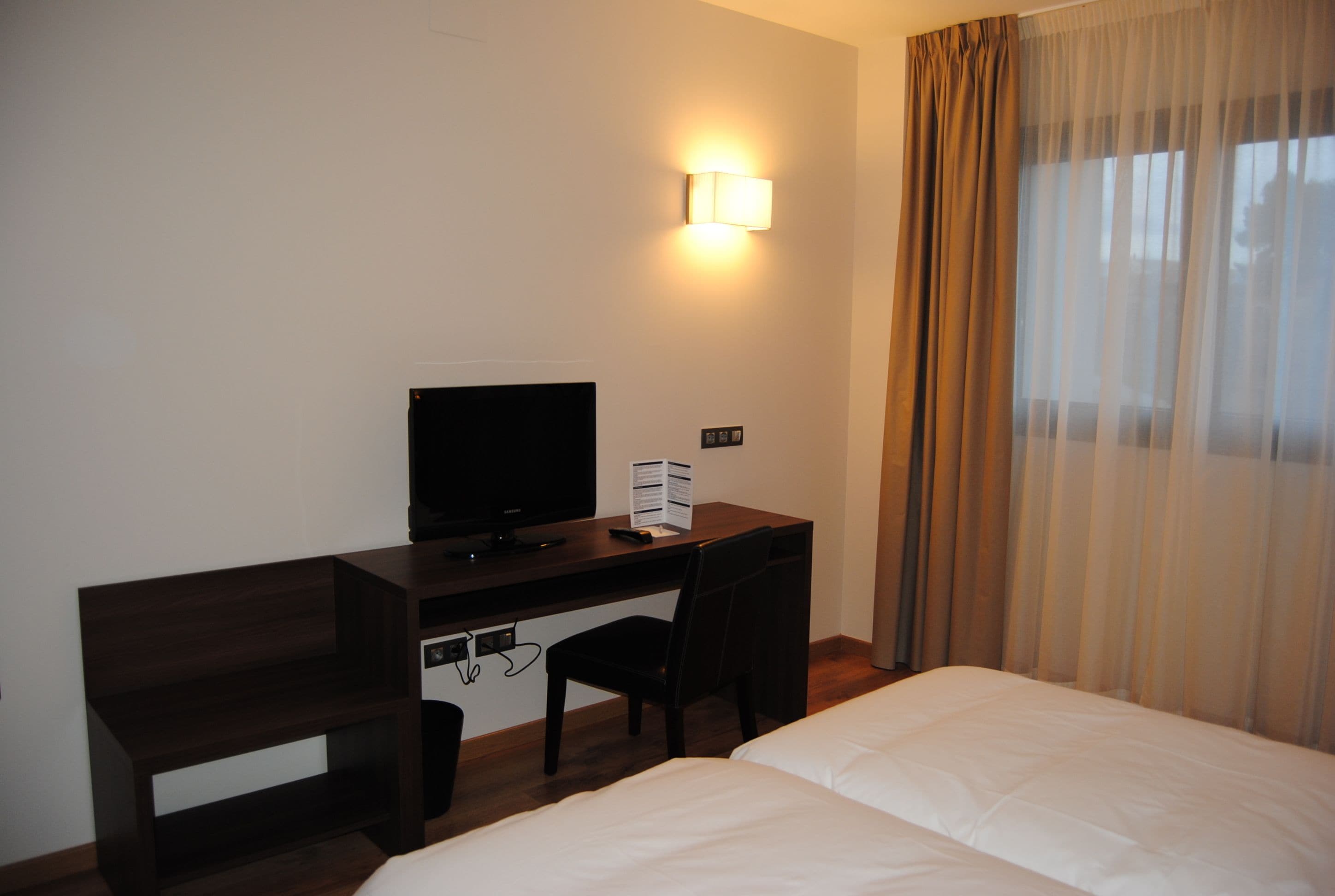 A hotel room with a bed, a desk with a television, a chair, and a window with curtains.