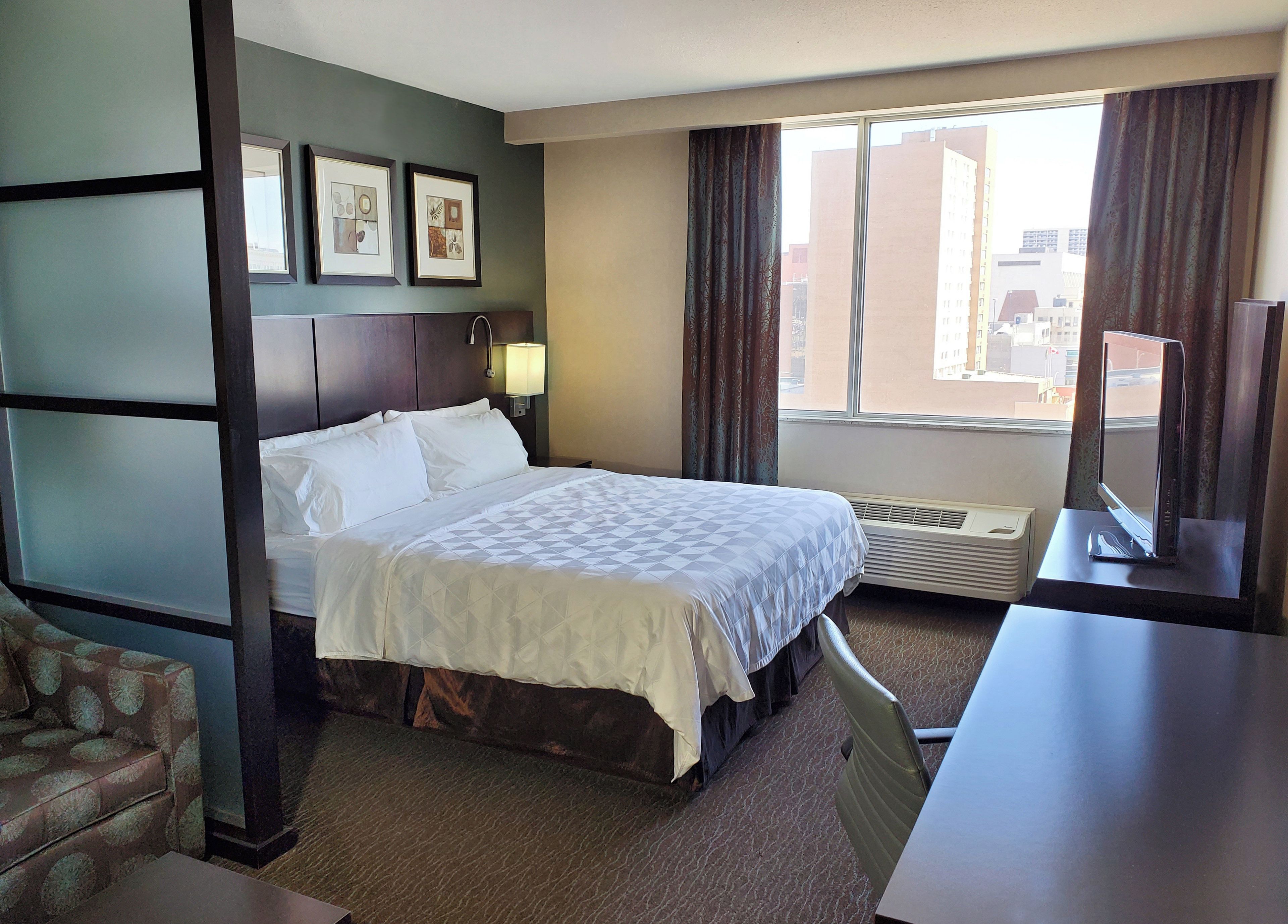 Een hotelkamer met een bed, bureau, stoel, raam met gordijnen, een aan de muur bevestigde televisie en ingelijste kunstwerken.
