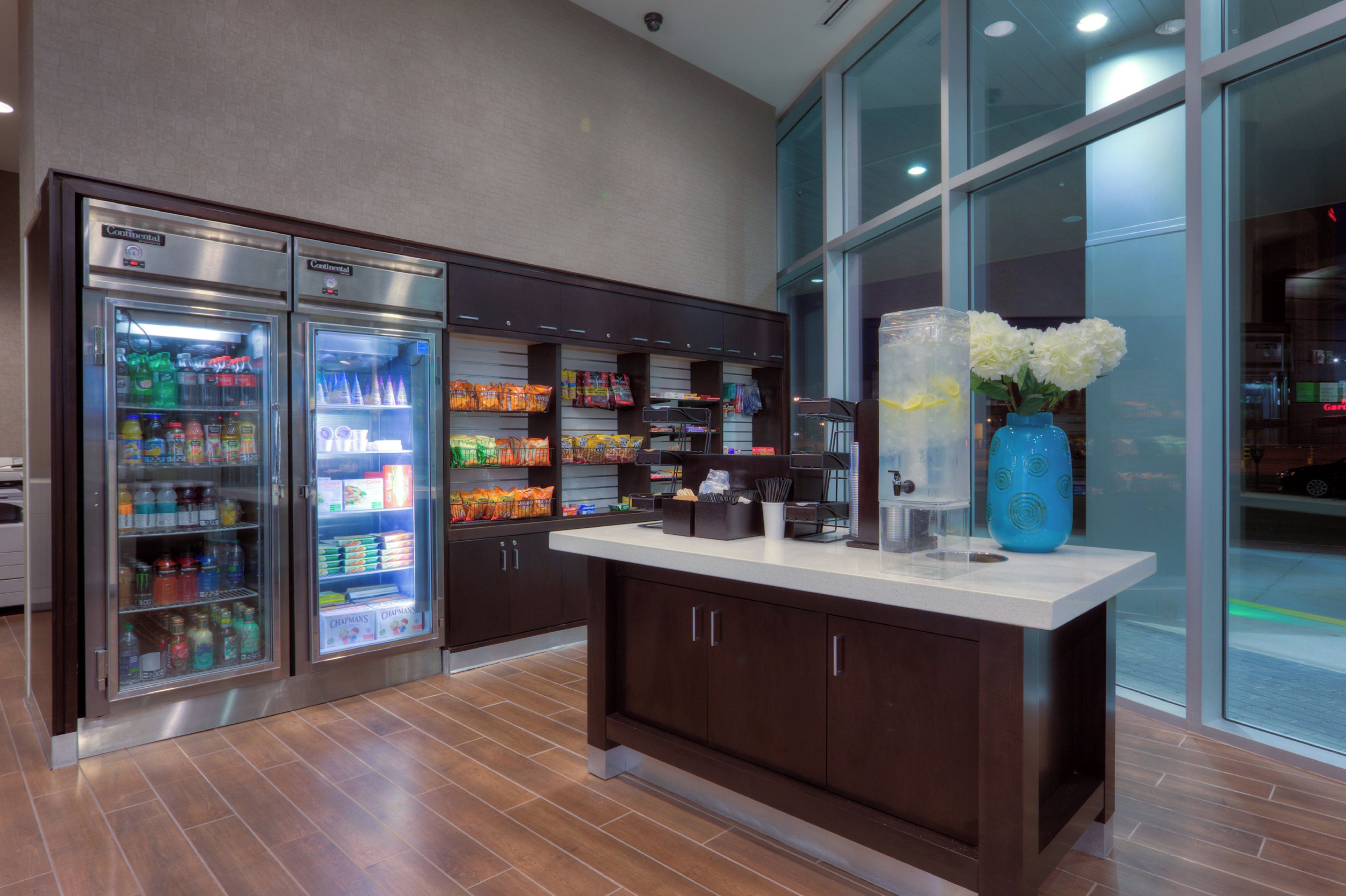 A commercial kitchen area with stainless steel refrigerators stocked with beverages, a counter with a vase of white flowers, and a large window providing a view of the outside.