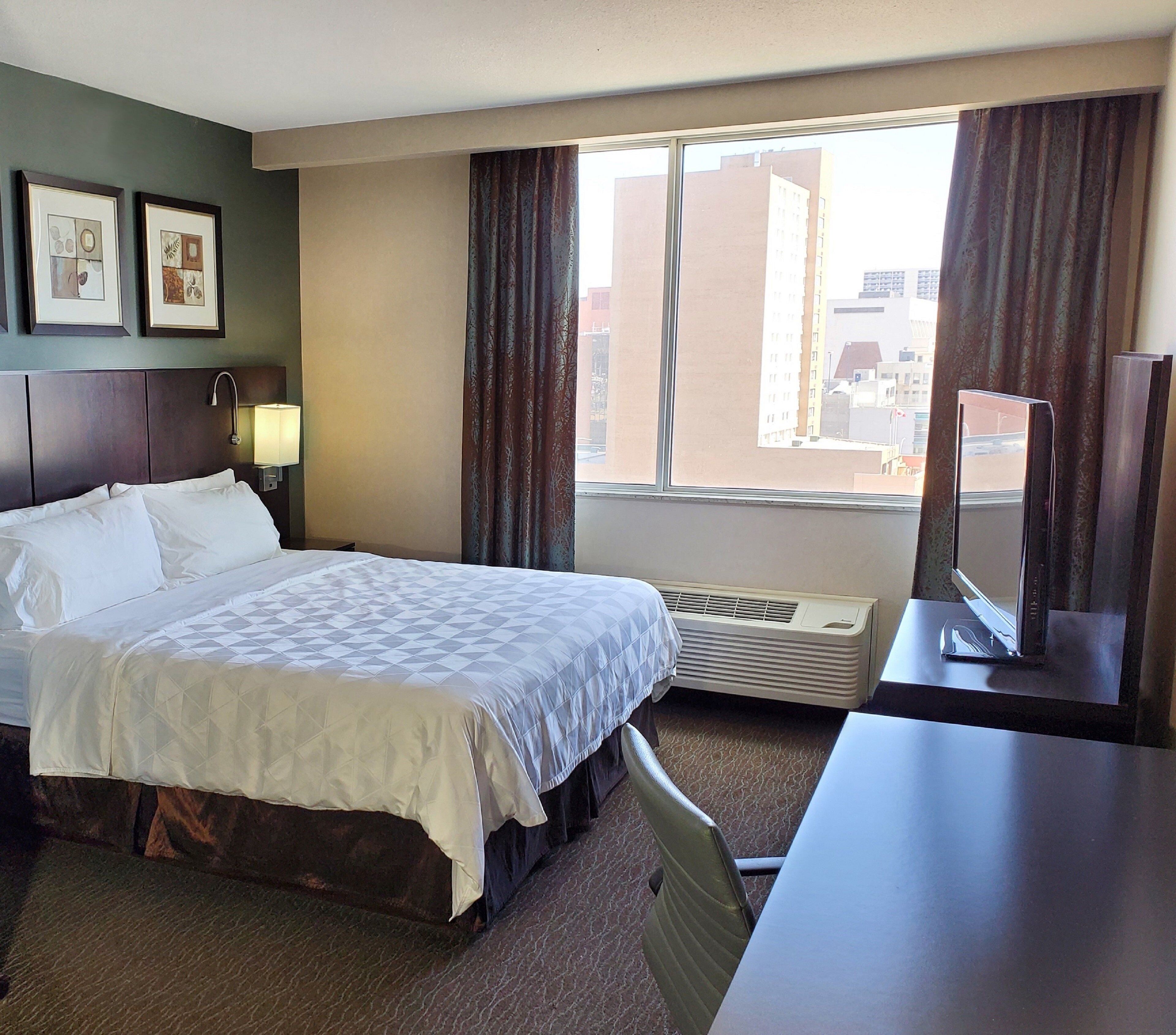 A hotel room with a bed, a desk, a TV, a window with curtains, and framed pictures on the wall.