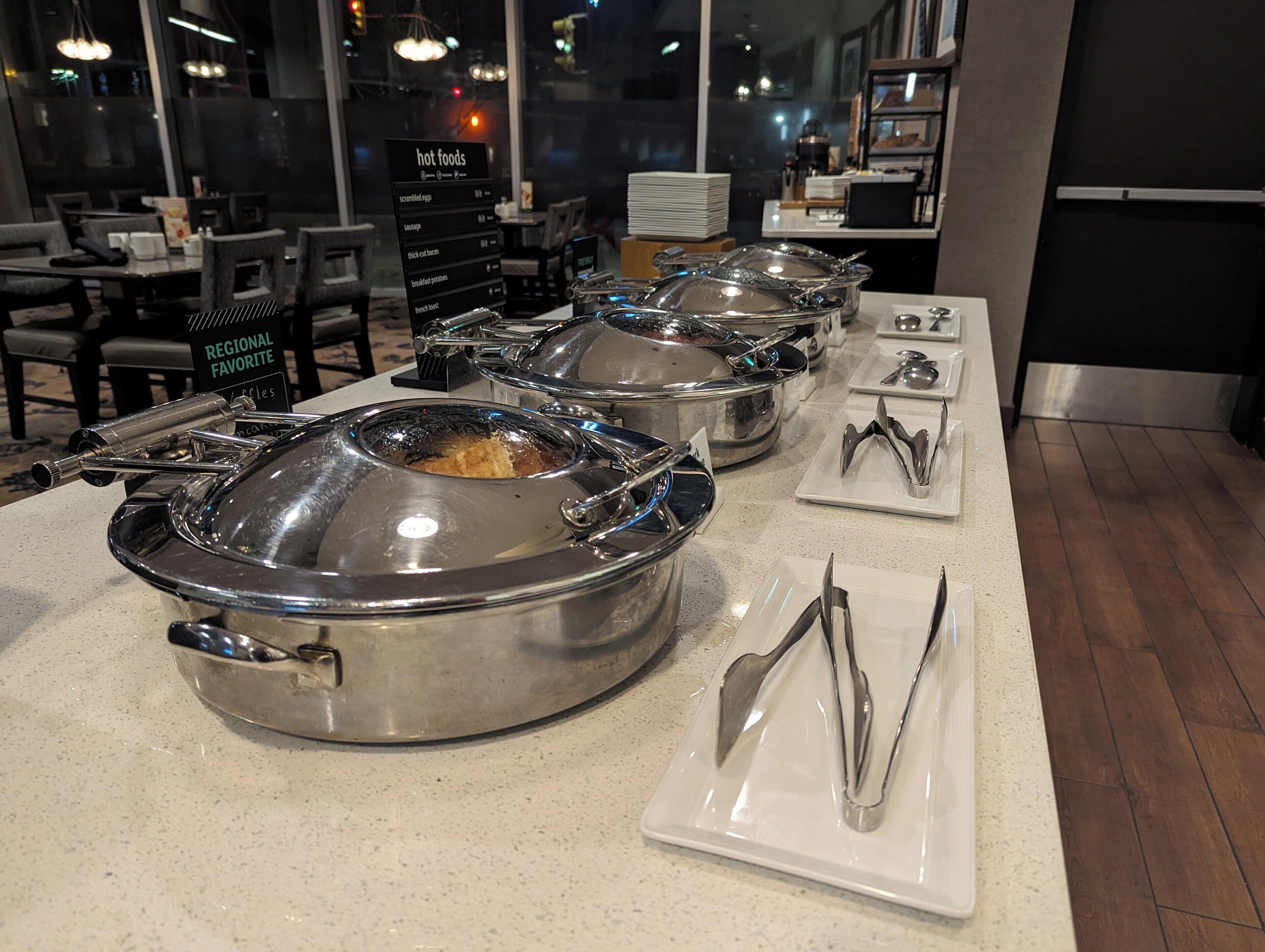 Estaciones de comida caliente tipo buffet, con utensilios para servir y un cartel que indica las especialidades de la región.