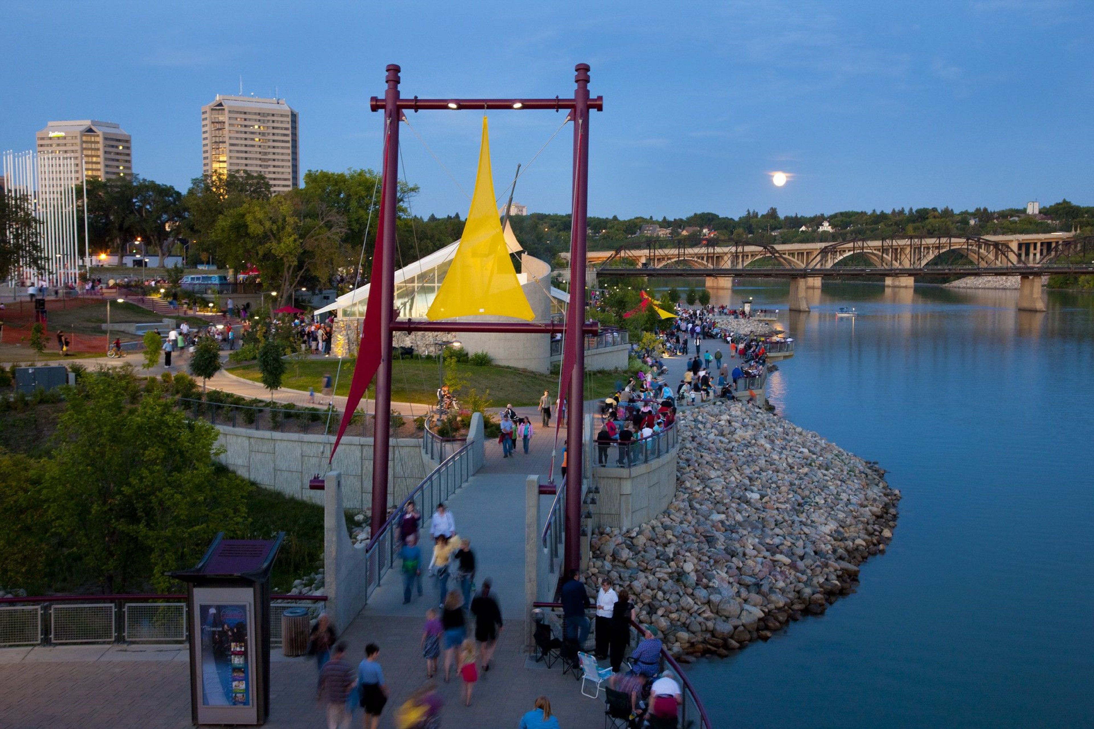 A riverside walkway with a yellow structure, a stone embankment, and a bridge in the background.