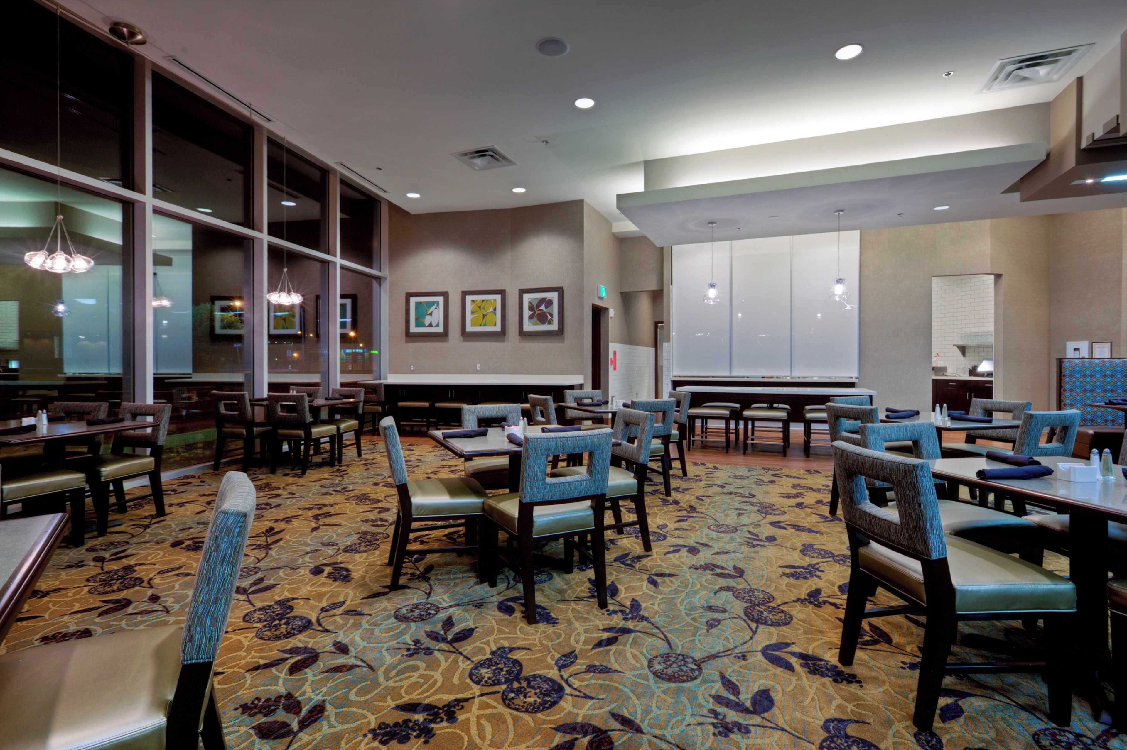 A hotel dining area with tables and chairs, large windows, and artwork on the walls.