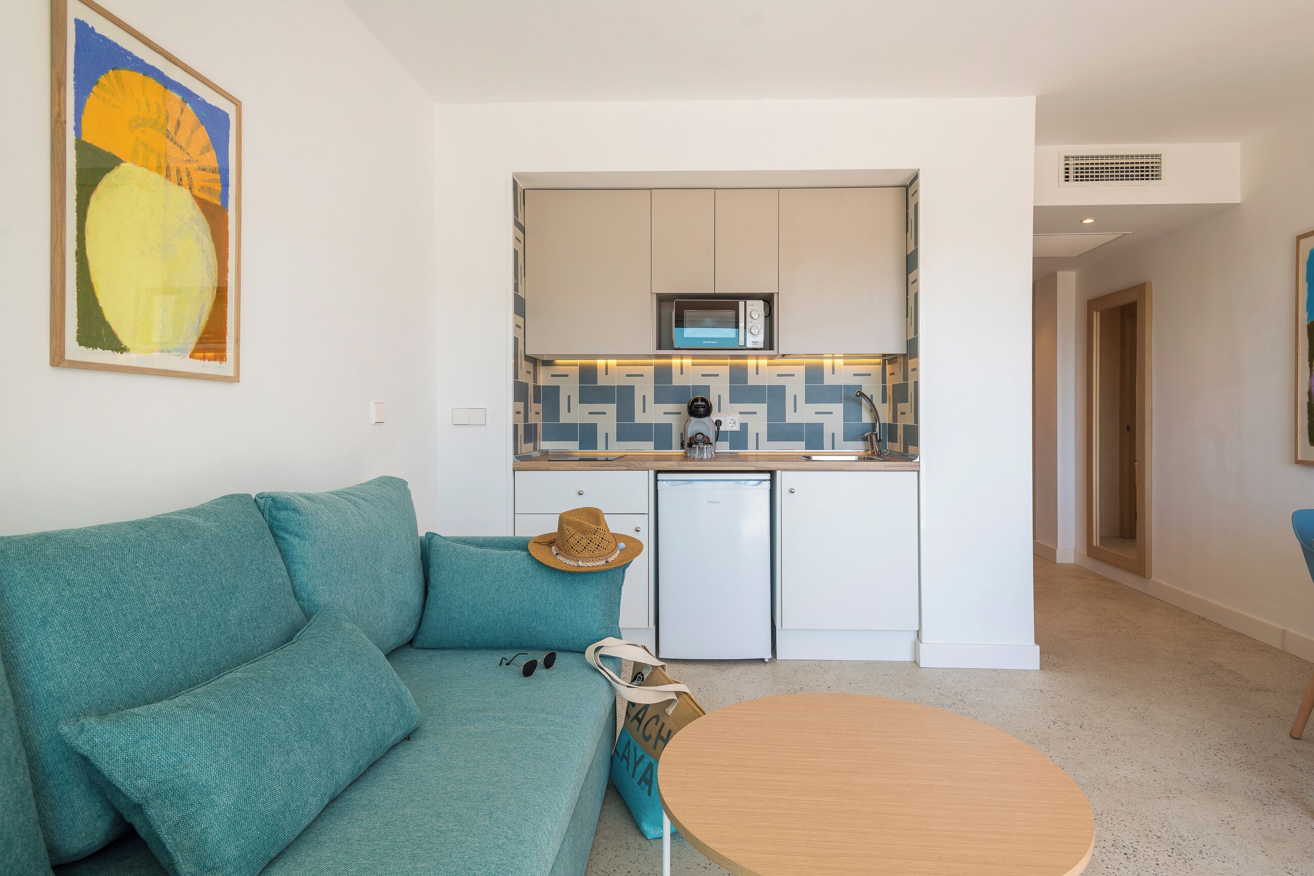 Un espace de vie compact comprenant un canapé de couleur turquoise, une petite table ronde, un coin cuisine avec des meubles blancs et un four à micro-ondes, ainsi qu’un pare-feu décoratif.
