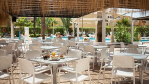 An outdoor dining area with white plastic chairs and tables, a pool in the background, and palm trees.