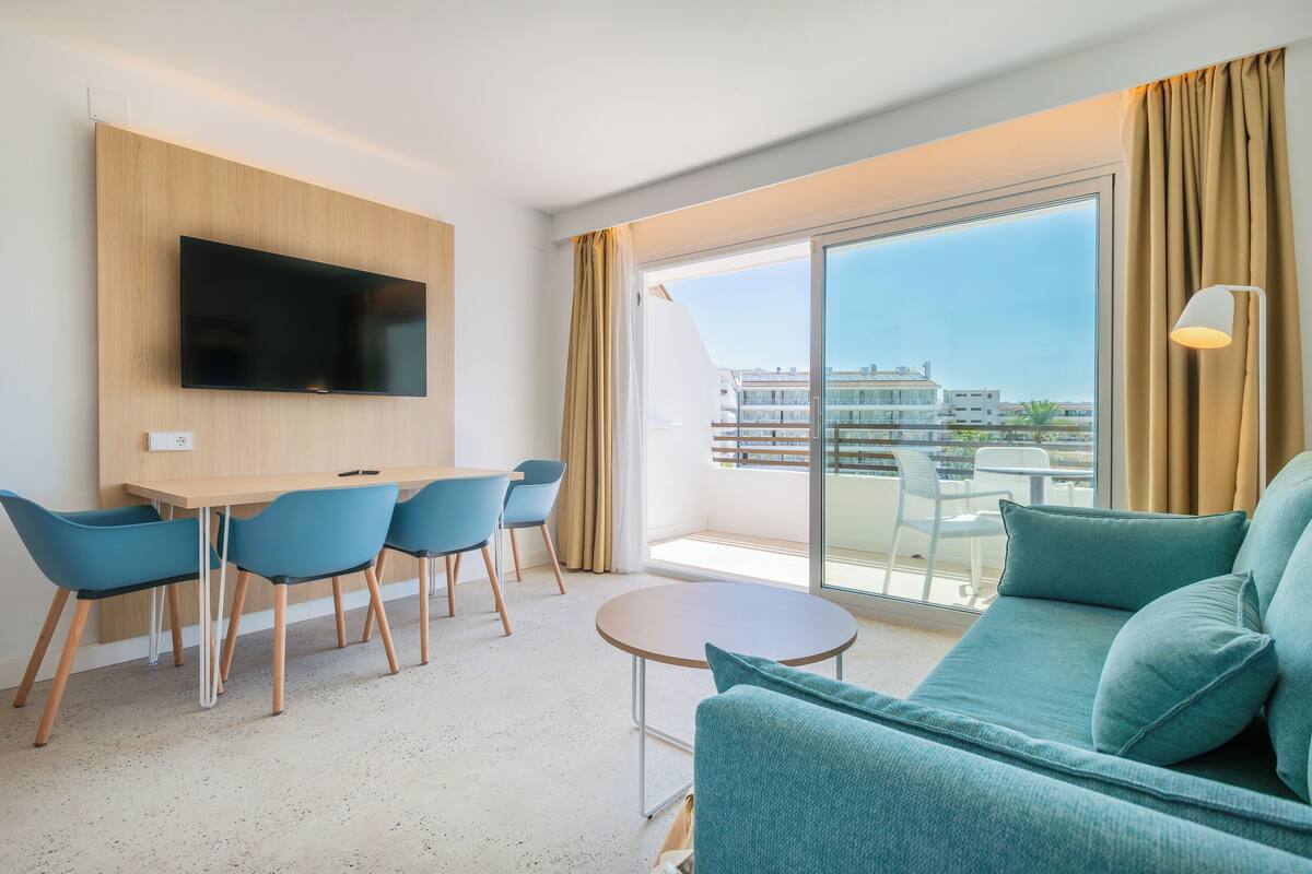 Une chambre d’hôtel moderne dotée d’une grande fenêtre, d’un téléviseur à écran plat fixé au mur, d’un canapé de couleur turquoise, d’une petite table ronde et d’un coin repas avec des chaises bleues.