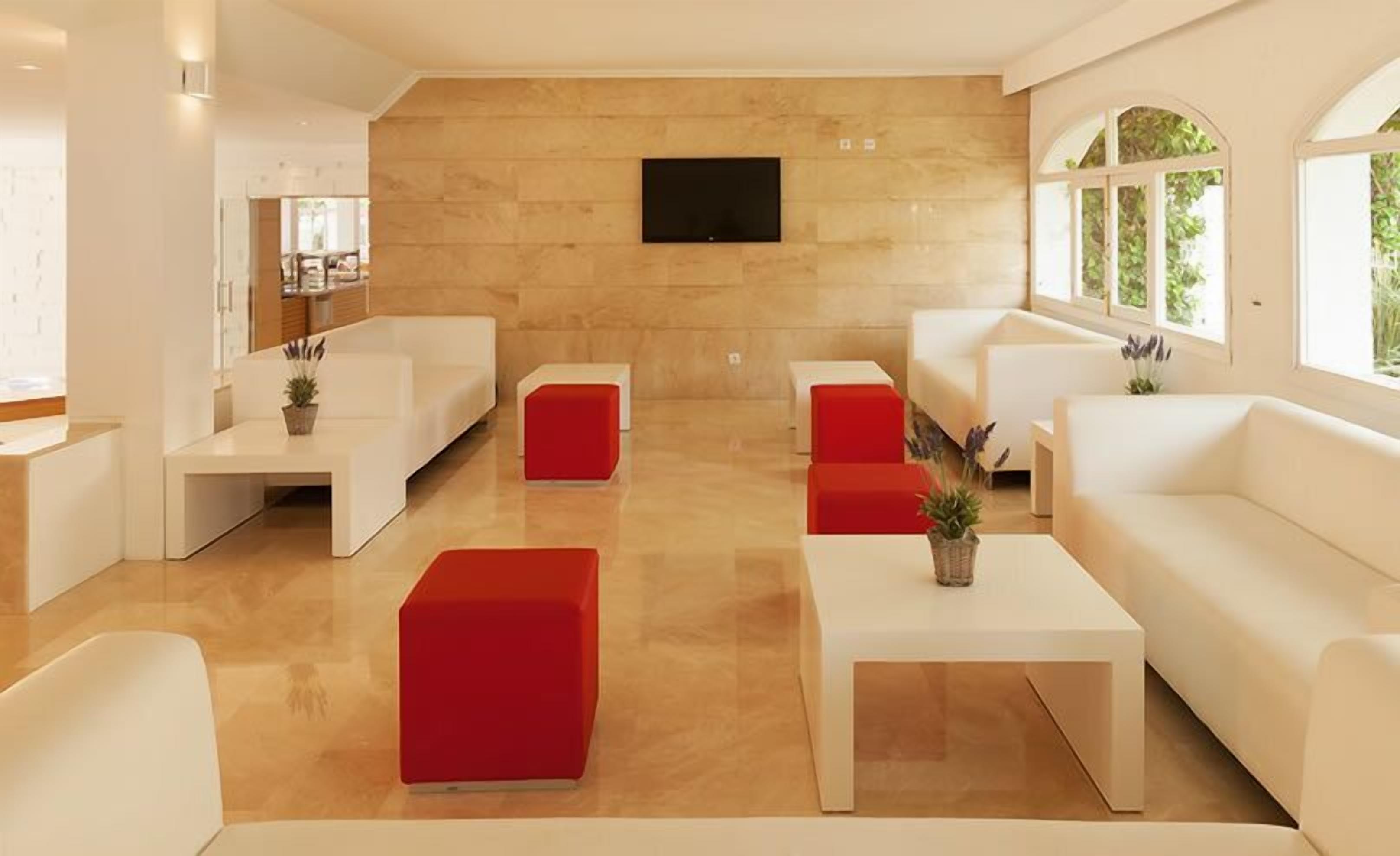 A modern living room with white sofas, red accent chairs, and a flat-screen TV mounted on a wooden wall.