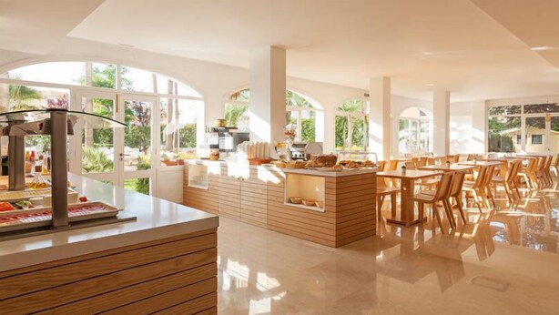 A modern kitchen with wooden cabinetry and a central island, large windows offering views of greenery, and a dining area with wooden tables and chairs.